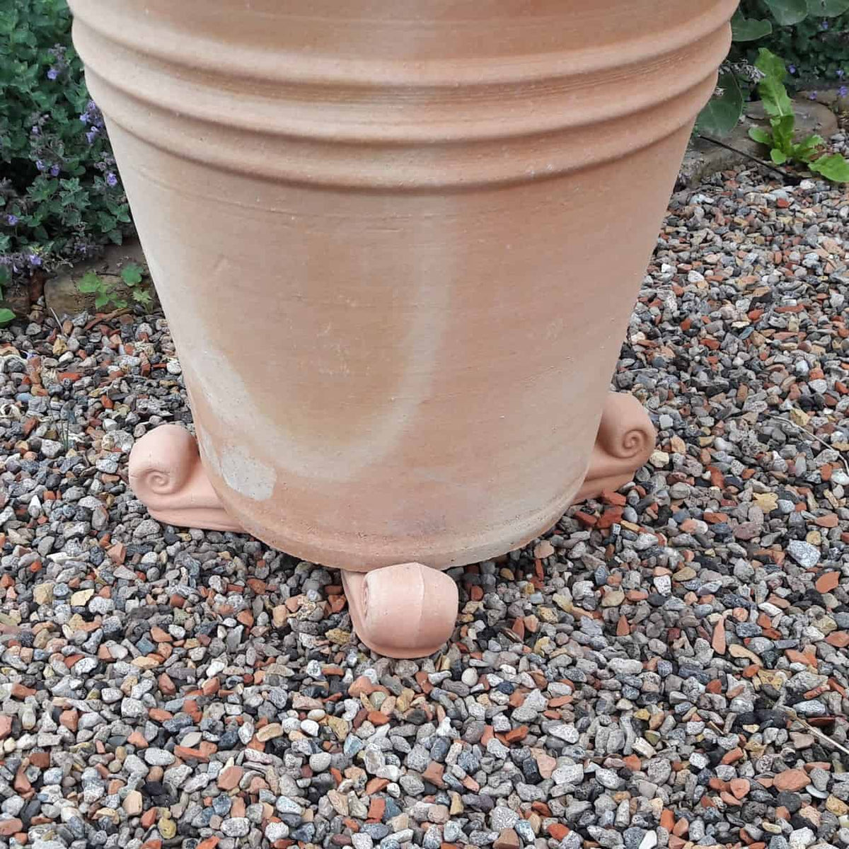 Ein großer Terrakotta-Pflanzkübel steht auf einer Kiesfläche, erhöht durch drei kleine, dekorative Stützen, die an stilisierte Tierfüße erinnern. Ein Terrakotta-Unterteller, Untersetzer 30 cm sorgt für die richtige Entwässerung. Im Hintergrund sind Pflanzen und Grünpflanzen teilweise sichtbar.
