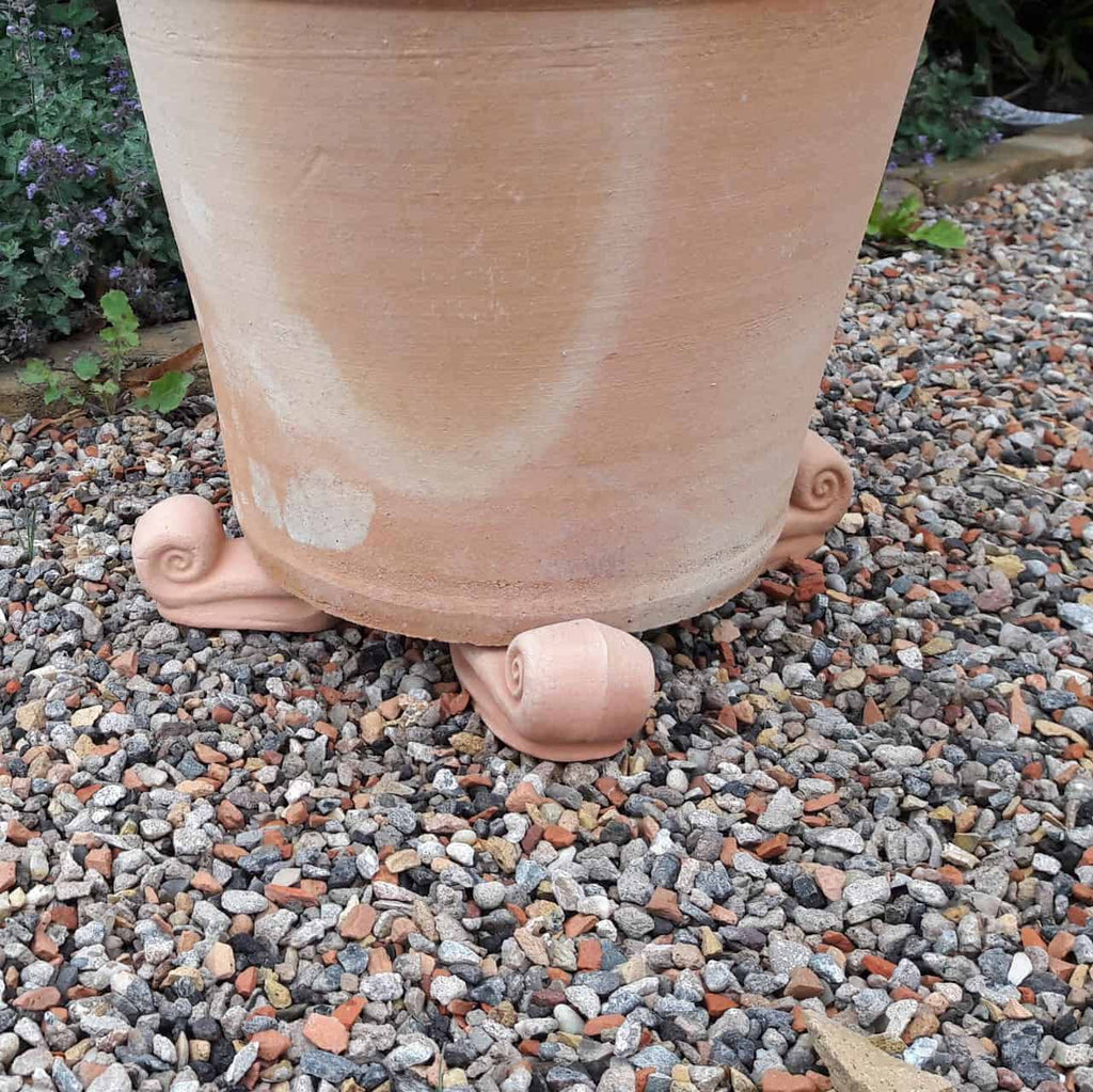 Ein großer Terrakottatopf steht auf einer Kiesfläche und wird von vier kleinen Terrakottafüßen in Spiralform erhöht. Darunter sorgt ein Terrakotta-Unterteller (Untersetzer 30 cm) für Stabilität und Entwässerung. Im Hintergrund sind grüne Pflanzen zu sehen.
