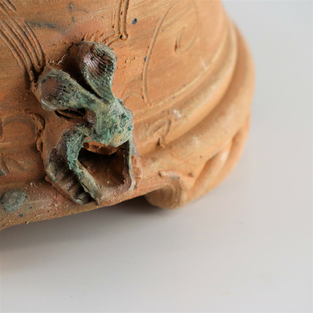 Nahaufnahme des handgefertigten Pflanzgefäßes aus Terrakotta, quadratisch, mit einem Griff in Form einer verdrehten, verwitterten Schleife, die aus einer rauen, strukturierten Oberfläche ragt. Die Keramik hat eine erdbraune Farbe mit subtilen grünlichen Patina-Akzenten, die auf Alter und Gebrauch hinweisen.