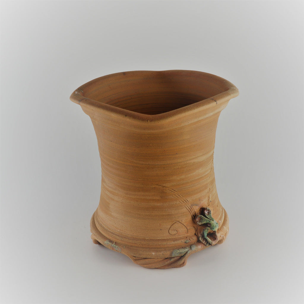Das **handgefertigte Pflanzgefäß aus Terracotta, Quadratisch** hat einen breiten, geschwungenen Rand und einen leicht konischen Boden. Der Topf ist am Boden mit einem filigranen kleinen Ranken- und Blumendetail verziert, das seinem schlichten Design einen Hauch natürlicher Eleganz verleiht. Der Hintergrund ist in einem weichen, neutralen Farbton gehalten.