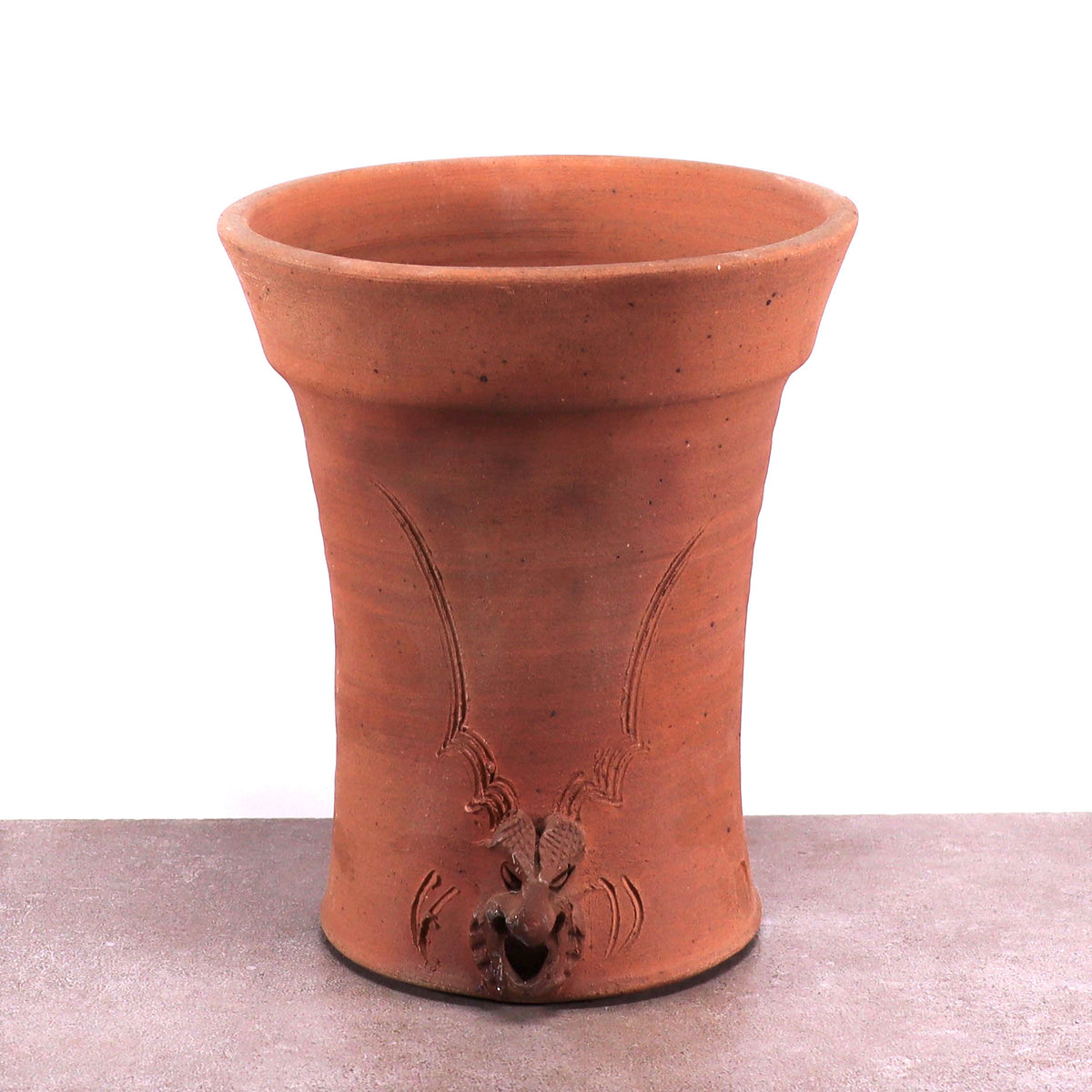 Ein hoher, rotbrauner, frostfester Blumentopf aus Terrakotta mit ausgestelltem Rand und glatter Oberfläche zeigt ein geformtes dekoratives Motiv mit dem Gesicht eines geweihten Wesens. Der schlichte weiße Hintergrund lässt den Blumentopf auf einer flachen, grauen Oberfläche hervorstechen.