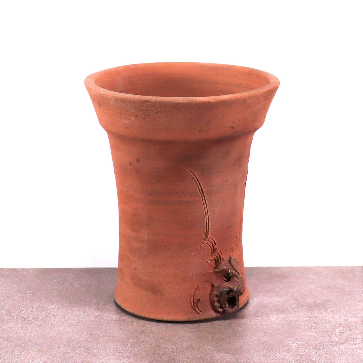 Ein rustikaler, zylindrischer Blumentopf aus frostfester Terrakotta mit breitem, ausgestelltem Rand und strukturiertem Drachenrelief in Bodennähe, ruht auf einer grauen Oberfläche vor weißem Hintergrund.