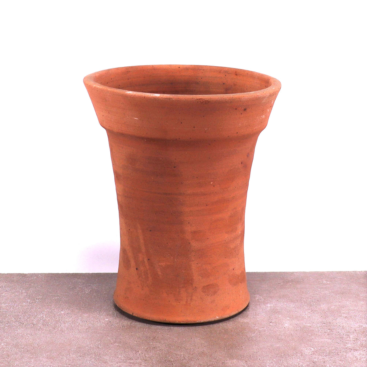 Ein einzelner frostfester Terrakotta-Blumentopf mit leicht ausgestelltem Rand steht auf einer flachen, grauen Oberfläche vor weißem Hintergrund. Der Topf hat eine glatte, aber subtil strukturierte Oberfläche und weist die erdige, rötlich-braune Farbe auf, die für Terrakotta charakteristisch ist.