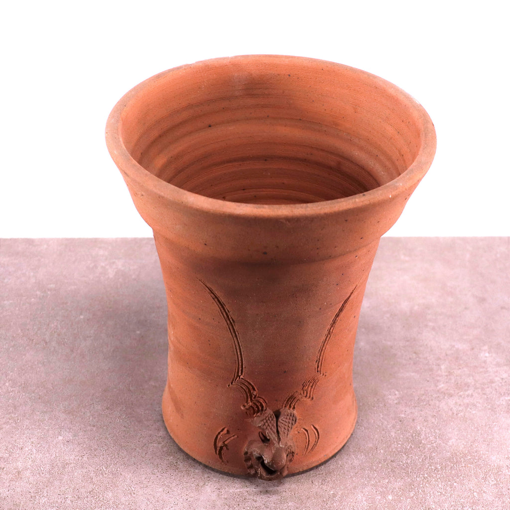Ein handgefertigter „frostfester Terrakotta-Blumentopf“ in einem satten Rotbraun mit ausgestelltem Rand und aufwendigen dekorativen Schnitzereien an der Außenseite. In der Nähe des Bodens befindet sich eine kleine Tierfigur, und der Topf steht auf einer strukturierten Oberfläche vor einem schlichten weißen Hintergrund.
