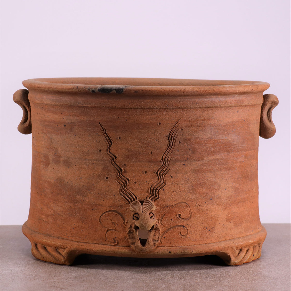 Der frostfeste, ovale Pflanztopf aus Terrakotta ist ein großer Pflanztopf mit rustikalem Finish, zwei seitlichen Griffen und einem geprägten Design eines gehörnten Tiergesichts mit verzierten, fließenden Hörnern auf der Vorderseite. Der Sockel weist ein geschnitztes Muster auf und steht auf einer glatten Oberfläche.