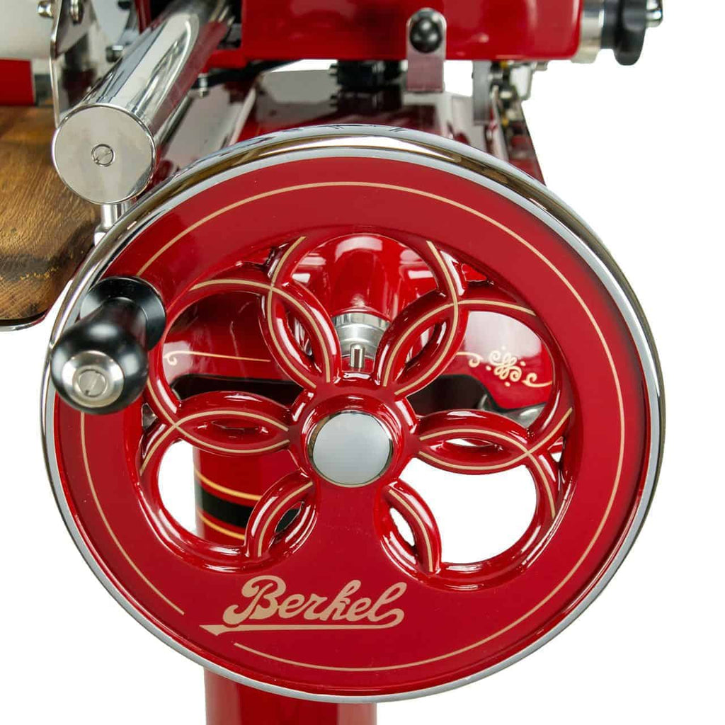 Nahaufnahme eines Vintage-Schneidebretts vom Typ Berkel Volano Tribute Rot mit einer verzierten Handkurbel mit dekorativem Blumenmuster. Der Name „Berkel“ ist in eleganter Schrift deutlich auf der Oberfläche des Rads zu sehen, ergänzt durch Ständer und Schneidebrett aus Fassholz.