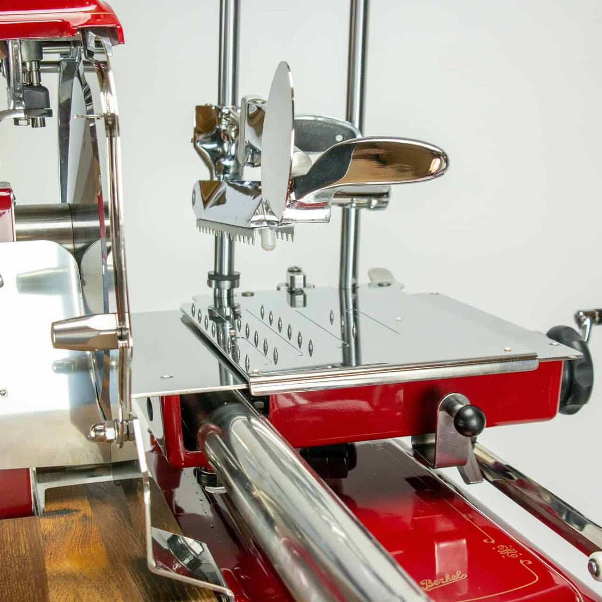 Eine Nahaufnahme einer Berkel Volano Tribute Rot Aufschnittmaschine mit Blumenschwungrad mit Ständer und Schneidebrett aus Fassholz. Die Maschine besticht durch eine glänzende Metalloberfläche mit auffälligen roten Akzenten. Schneideblatt, Schlitten und Bedienelemente sind deutlich zu erkennen und zeigen das aufwendige Design und die außergewöhnliche Handwerkskunst. Der Hintergrund ist schlicht und hell.