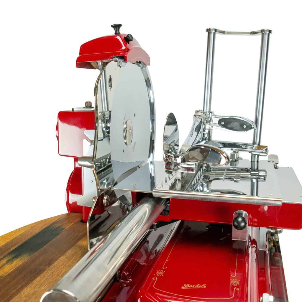 Auf einer Holzoberfläche wird ein Berkel Volano Tribute Rot-Fleischschneider im rot-silbernen Vintage-Design mit runder Klinge, Griff, verschiedenen Chromkomponenten und einem schönen Schwungrad mit Blumenmuster mit Ständer und Schneidebrett aus Fassholz präsentiert. Die Maschine zeigt eine aufwendig gestaltete Retro-Ästhetik mit sorgfältig gefertigten Metall- und Mechanikteilen.