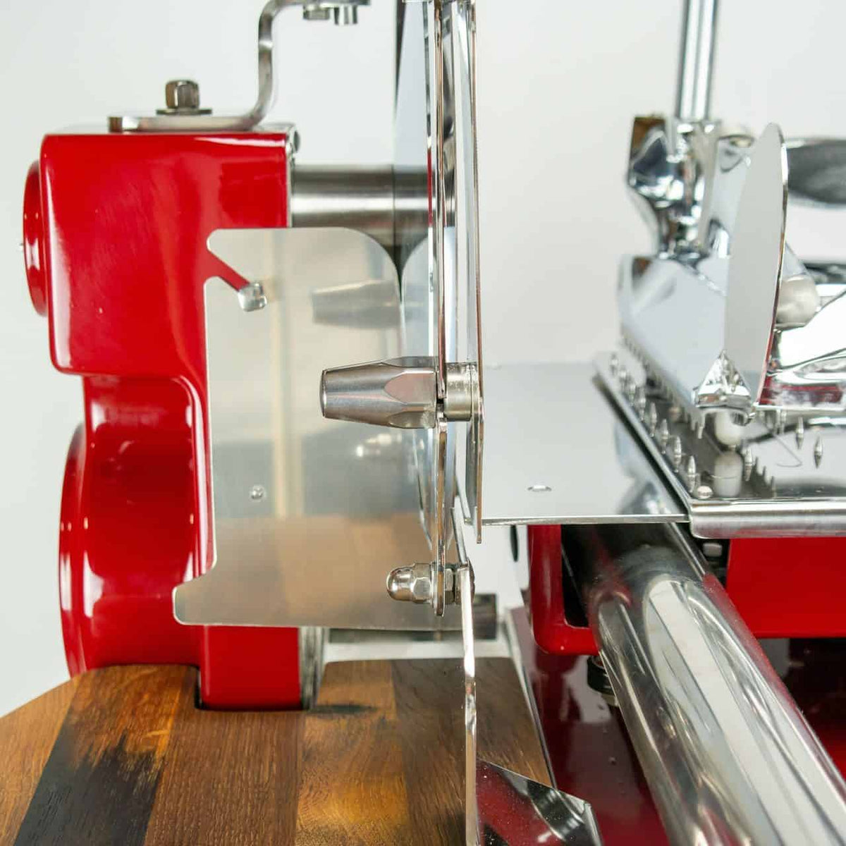 Eine Nahaufnahme eines Berkel Volano Tribute Rot-Fleischschneiders mit floralem Schwungrad, platziert auf einem aus Fassholz gefertigten Schneidebrett. Der rot-silberne Allesschneider weist eine runde Klinge, einen Einstellgriff und verschiedene Metallkomponenten auf. Die Klinge ist teilweise mit einem Schutzschild bedeckt.