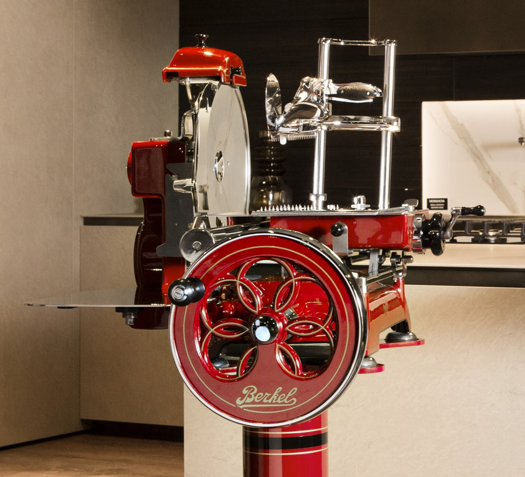 Eine Berkel Volano Tribute Rot Aufschnittmaschine mit Blumenschwungrad und Aufschnittbrett aus Weinfassholz steht auf einem passenden Sockel in einer modernen Küche. Der Allesschneider verfügt über Chromakzente, eine große kreisförmige Klinge und einen Blumenschwungrad mit Handkurbel und aufwendigen Details. Der Hintergrund zeigt eine beige Arbeitsplatte und dunkle Schränke.