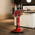 Ein Vintage-Fleischschneider Berkel Volano Tribute Rot mit handbetriebenem Blumenschwungrad und Fassholz-Schneidebrett steht auf einem Podest in einer modernen Küche. Der Allesschneider verfügt über polierte Metallkomponenten und ein kompliziertes Design. Im Hintergrund ist ein Herd mit Töpfen auf einer hellen Arbeitsplatte zu sehen.