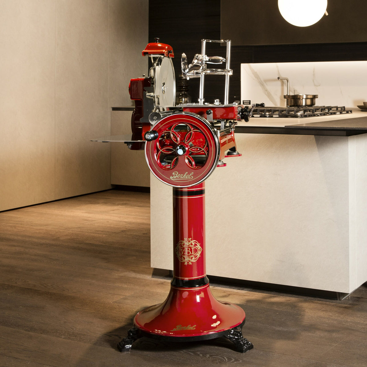 Ein Vintage-Fleischschneider Berkel Volano Tribute Rot mit handbetriebenem Blumenschwungrad und Fassholz-Schneidebrett steht auf einem Podest in einer modernen Küche. Der Allesschneider verfügt über polierte Metallkomponenten und ein kompliziertes Design. Im Hintergrund ist ein Herd mit Töpfen auf einer hellen Arbeitsplatte zu sehen.