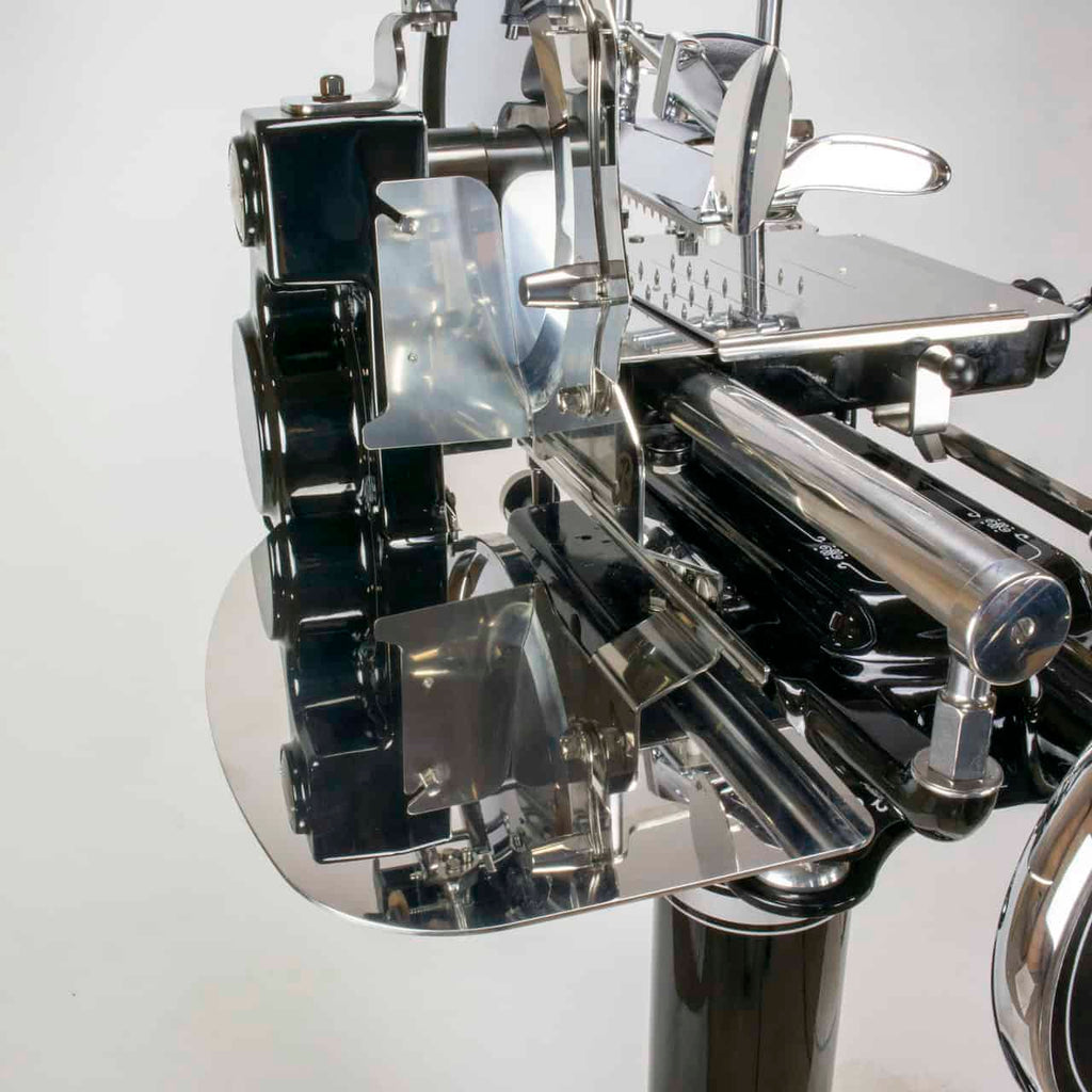 Nahaufnahme eines alten mechanischen Geräts mit verschiedenen Hebeln, Knöpfen und Metallkomponenten. Die Berkel Volano Tribute, schwarz, manuelle Aufschnittmaschine mit Blumenschwungrad + Standfuß + Aufschnittbrett aus Fassholz verfügt über eine glänzende, reflektierende Oberfläche und komplizierte Teile, die an eine alte Schreibmaschine oder Industriemaschine erinnern. Der Hintergrund ist schlicht und weiß.