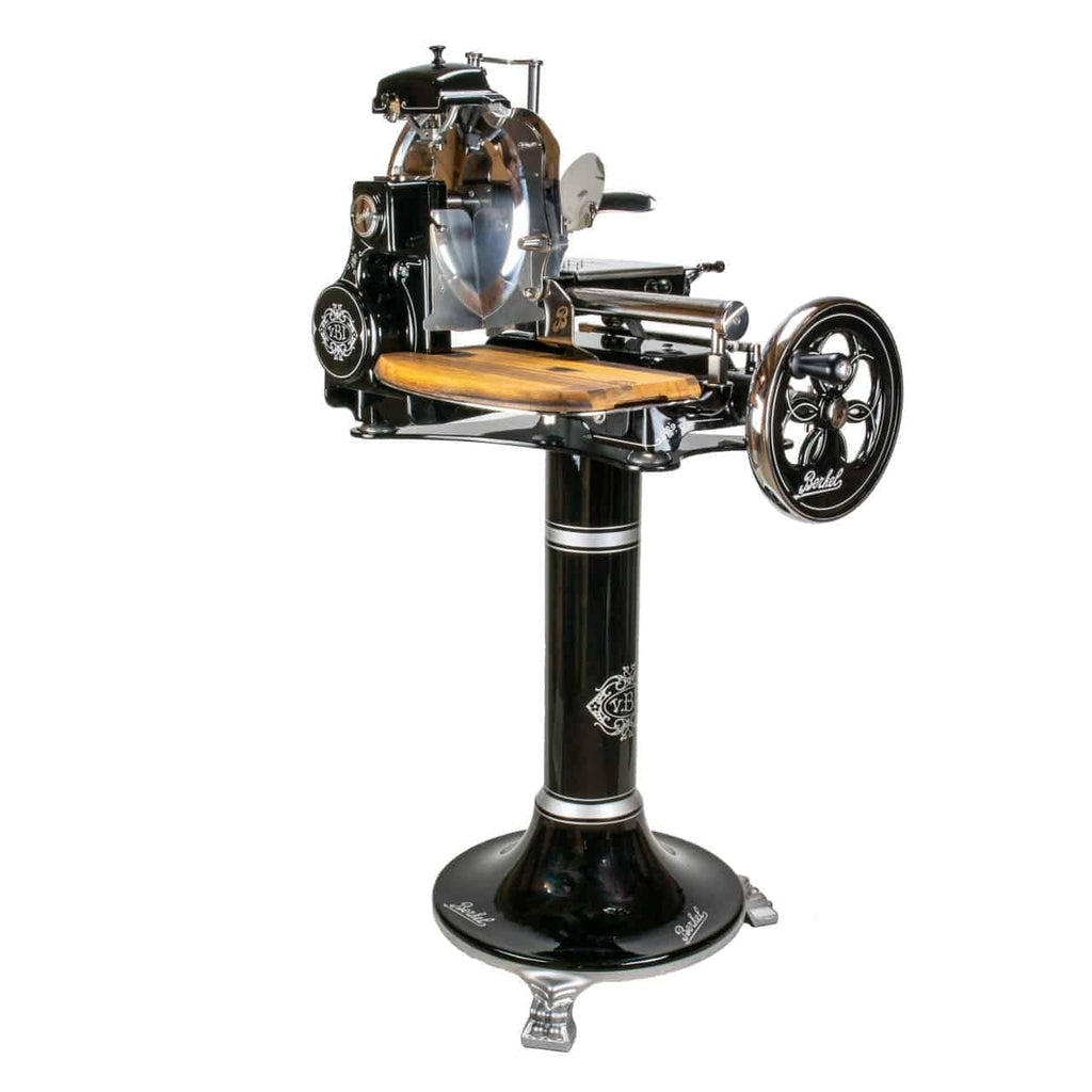 Ein Berkel Volano Tribute, schwarzer antiker Fleischschneider mit aufwendigen Verzierungen, auf einem Sockel montiert. Er verfügt über ein hölzernes Schneidebrett, ein großes Blumenschwungrad (Handkurbelrad) und polierte Metallteile. Der Sockel, ebenfalls schwarz, zeigt dekorative Elemente und den Markennamen „Berkel“.