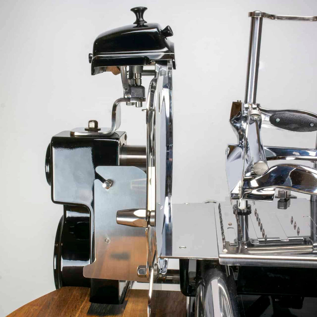 Eine Nahaufnahme der manuellen Aufschnittmaschine Berkel Volano Tribute, schwarz, mit Blumenschwungrad + Standfuß + Aufschnittbrett aus Fassholz. Der Allesschneider hat ein glänzendes Metallgehäuse mit schwarzen Akzenten, eine sichtbare Klinge und einen Griff. Er steht auf einer Holzoberfläche aus Fassholz und verfügt über Komponenten wie einen Anspitzer, ein Motorgehäuse und ein Blumenschwungrad vor einem schlichten weißen Hintergrund.