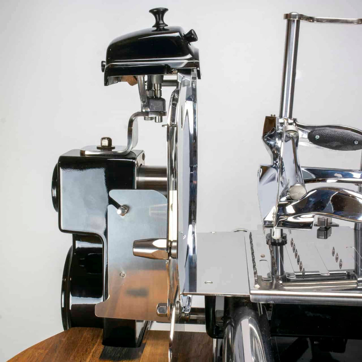 Eine Nahaufnahme der manuellen Aufschnittmaschine Berkel Volano Tribute, schwarz, mit Blumenschwungrad + Standfuß + Aufschnittbrett aus Fassholz. Der Allesschneider hat ein glänzendes Metallgehäuse mit schwarzen Akzenten, eine sichtbare Klinge und einen Griff. Er steht auf einer Holzoberfläche aus Fassholz und verfügt über Komponenten wie einen Anspitzer, ein Motorgehäuse und ein Blumenschwungrad vor einem schlichten weißen Hintergrund.