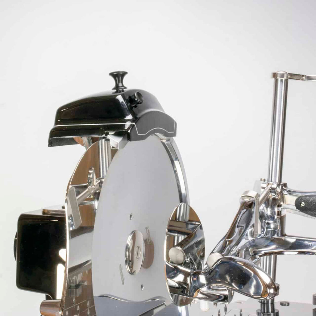 Eine Nahaufnahme eines alten mechanischen Fleischschneiders, genauer gesagt des Berkel Volano Tribute. Der Schneider verfügt über eine große, runde Klinge und glänzende Metallkomponenten. Oben ist ein schwarzer Griff sichtbar. Der Hintergrund ist schlicht und weiß und unterstreicht die Eleganz dieser schwarzen manuellen Aufschnittmaschine mit Blumenschwungrad + Standfuß + Aufschnittbrett aus Fassholz.