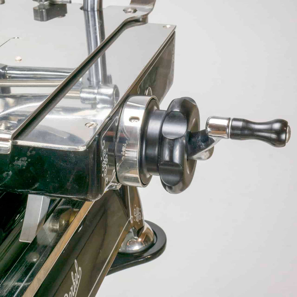 Nahaufnahme einer manuell betriebenen Kurbel, die an der polierten, metallischen Aufschnittmaschine Berkel Volano Tribute angebracht ist. Der schwarze Griff weist silberne Akzente auf und die gesamte schwarze manuelle Aufschnittmaschine mit Blumenschwungrad + Standfuß + Aufschnittbrett aus Fassholz wirkt robust und industriell mit präzisen Details. Der Hintergrund ist neutral.