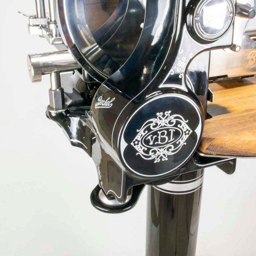 Nahaufnahme einer schwarzen, manuellen Fleischschneidemaschine „Berkel Volano Tribute“ im Vintage-Stil mit verzierten weißen Details und einer Schneidefläche aus Holz. Die Maschine verfügt über einen Handkurbelmechanismus und polierte Metallkomponenten. Auf der Maschine sind die Marke „Berkel“ und die Initialen „yBI“ zu sehen.