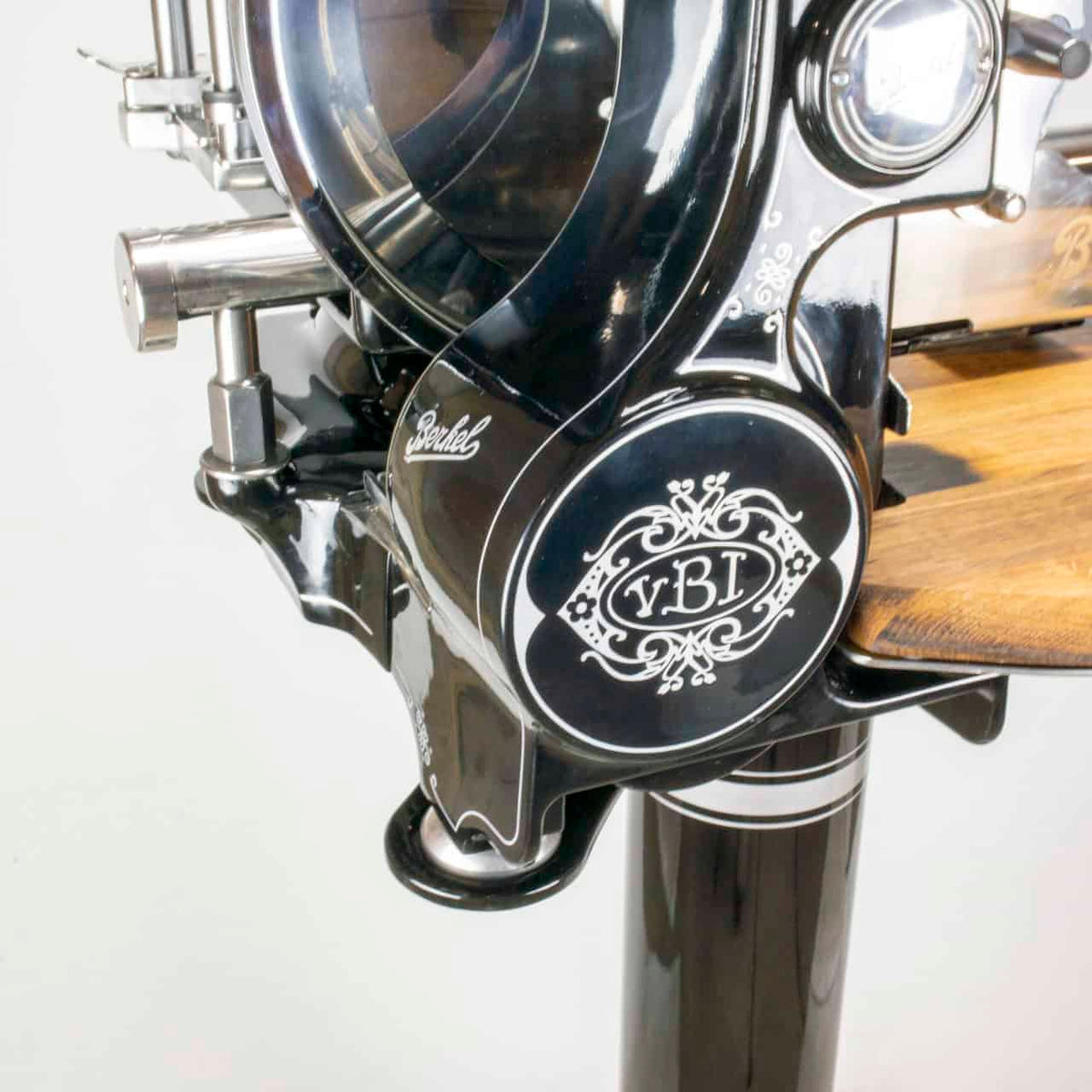 Nahaufnahme einer schwarzen, manuellen Fleischschneidemaschine „Berkel Volano Tribute“ im Vintage-Stil mit verzierten weißen Details und einer Schneidefläche aus Holz. Die Maschine verfügt über einen Handkurbelmechanismus und polierte Metallkomponenten. Auf der Maschine sind die Marke „Berkel“ und die Initialen „yBI“ zu sehen.