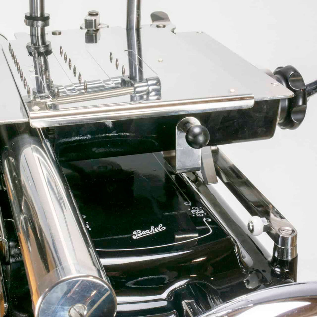 Eine Nahaufnahme der Berkel Volano Tribute, schwarz, manuellen Aufschnittmaschine mit Blumenschwungrad + Standfuß + Aufschnittbrett aus Fassholz offenbart ihren Vintage-Charme. Diese klassische Maschine verfügt über glänzende Metallkomponenten, darunter ein großes Rundmesser, einen Griff und eine flache Oberfläche für das Fleisch. Ihr glattes, poliertes Erscheinungsbild wird durch aufwendig gestaltete mechanische Details unterstrichen.