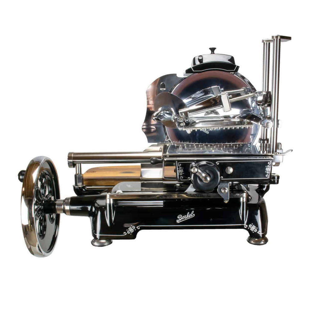 Der Berkel Volano Tribute Allesschneider in Schwarz ist ein Vintage-Fleischschneider mit Handbetrieb, der über ein großes rundes Messer und ein Handkurbelrad verfügt. Dieses Modell besticht durch dekorative Akzente und ein kunstvolles, poliertes Retro-Erscheinungsbild mit Blumenschwungrad-Design, komplett mit Ständer und Schneidebrett aus Fassholz.