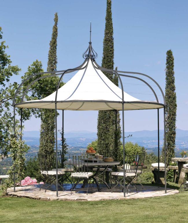 Ein charmanter Pavillon Solaire rund mit weißem Baldachin und schwarzem Metallrahmen steht auf einer Steinterrasse. Im Inneren gibt es einen Esstisch mit Stühlen, geschmückt mit einer Obstschale und Topfpflanzen. Hohe Bäume und eine malerische Hügellandschaft erstrecken sich unter einem klaren Himmel in die Ferne.
