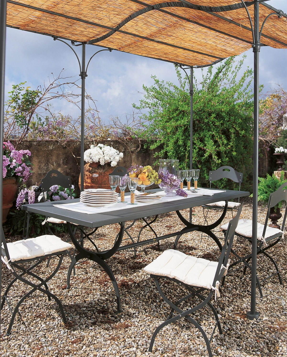Ein stilvolles Esszimmer im Freien besteht aus einem Tisch mit weißer Tischwäsche, Blumendekorationen und mehreren Gläsern unter einer freistehenden, gebogenen Pergola Solaire mit geflochtenem Baldachin. Umgeben von vier gepolsterten Stühlen ist der Terrassenbereich mit blühenden Blumen und Grünpflanzen geschmückt.