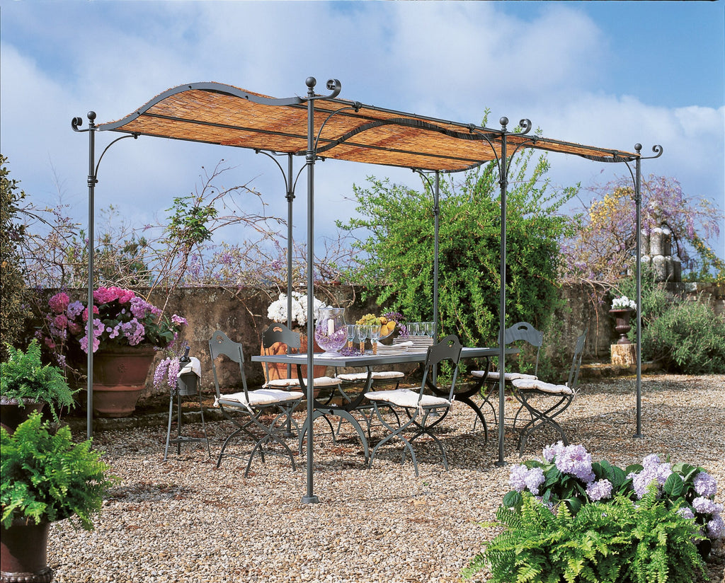 Eine bezaubernde Außenterrasse verfügt über ein schmiedeeisernes Esszimmerset mit sechs Stühlen und einem passenden Tisch unter einer freistehenden, gebogenen Pergola Solaire mit Strohdach. Die Terrasse ist von verschiedenen Topfpflanzen und blühenden Blumen umgeben, mit einer Steinmauer und üppigem Grün im Hintergrund.