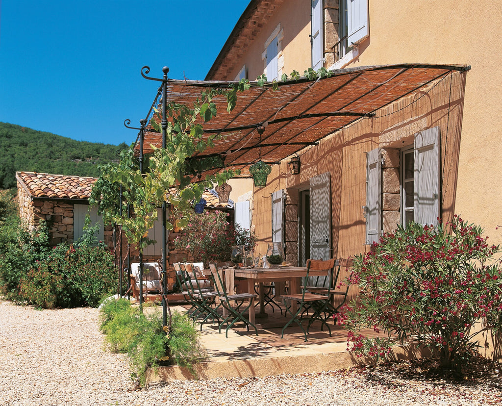 Eine gemütliche Außenterrasse mit Holztisch und Stühlen befindet sich unter einer mit Weinreben bewachsenen, gebogenen Anbaupergola Solaire, die an ein rustikales Steinhaus mit blauen Fensterläden angebaut ist. Üppiges Grün und blühende Pflanzen umgeben den Bereich und der Himmel ist klar und blau.
