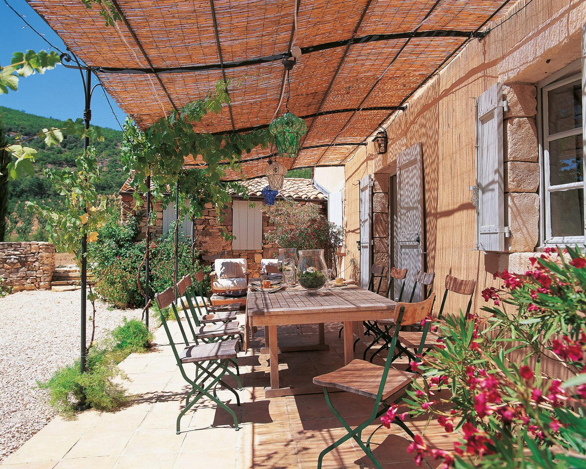 Ein rustikaler Essbereich im Freien mit einem Holztisch und grünen Metallstühlen wird von einer Gebogenen Anbaupergola Solaire beschattet. Die sonnenbeschienene Terrasse, geschmückt mit Hängepflanzen und umgeben von bunten Blumen und Steinmauern, bietet ebenfalls einen malerischen Blick ins Grüne, der durch die Eleganz der Solaire-Pergola noch unterstrichen wird.