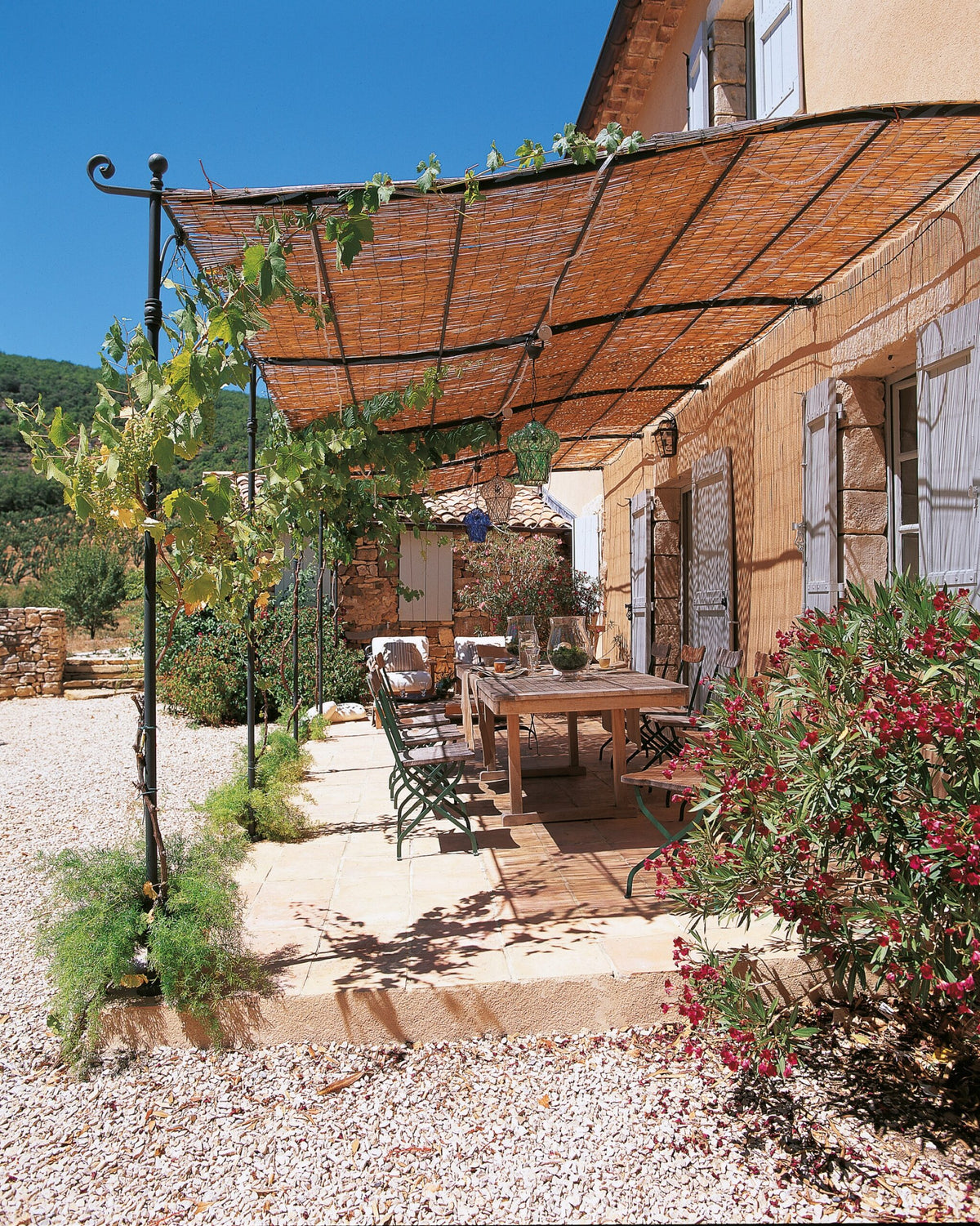 Eine sonnige Außenterrasse verfügt über eine Gebogene Anbaupergola Solaire, die mit Kletterpflanzen bedeckt ist. Darunter steht ein Holztisch mit Metallstühlen, alles zum Essen gedeckt. Die Terrasse grenzt an ein Steingebäude mit hellen Fensterläden und üppigen Büschen. Der Himmel darüber ist klar und blau.