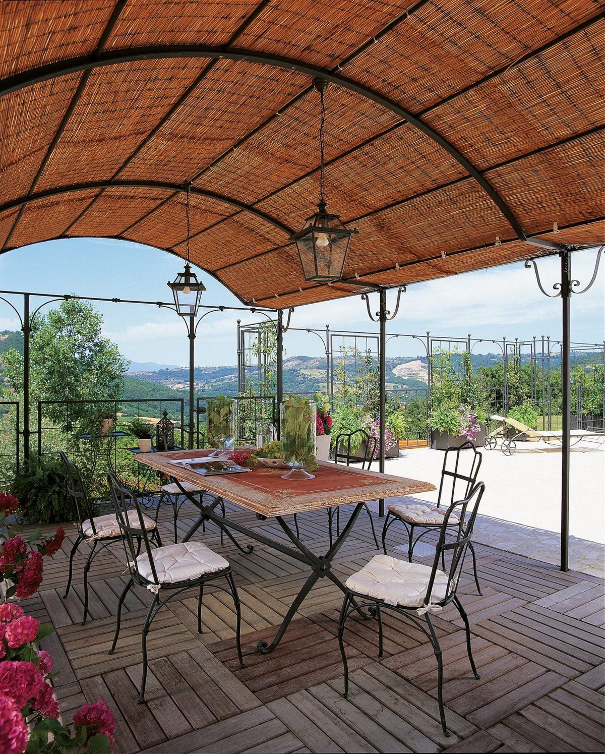 Eine schattige Außenterrasse mit einem Holztisch und Eisenstühlen mit weißen Kissen. Die Terrasse verfügt über eine lineare Anbaupergola Solaire mit geflochtenem Dach, Topfblumen und Panoramablick auf eine Hügellandschaft im Hintergrund. Laternenartige Lichter hängen von der Pergola.