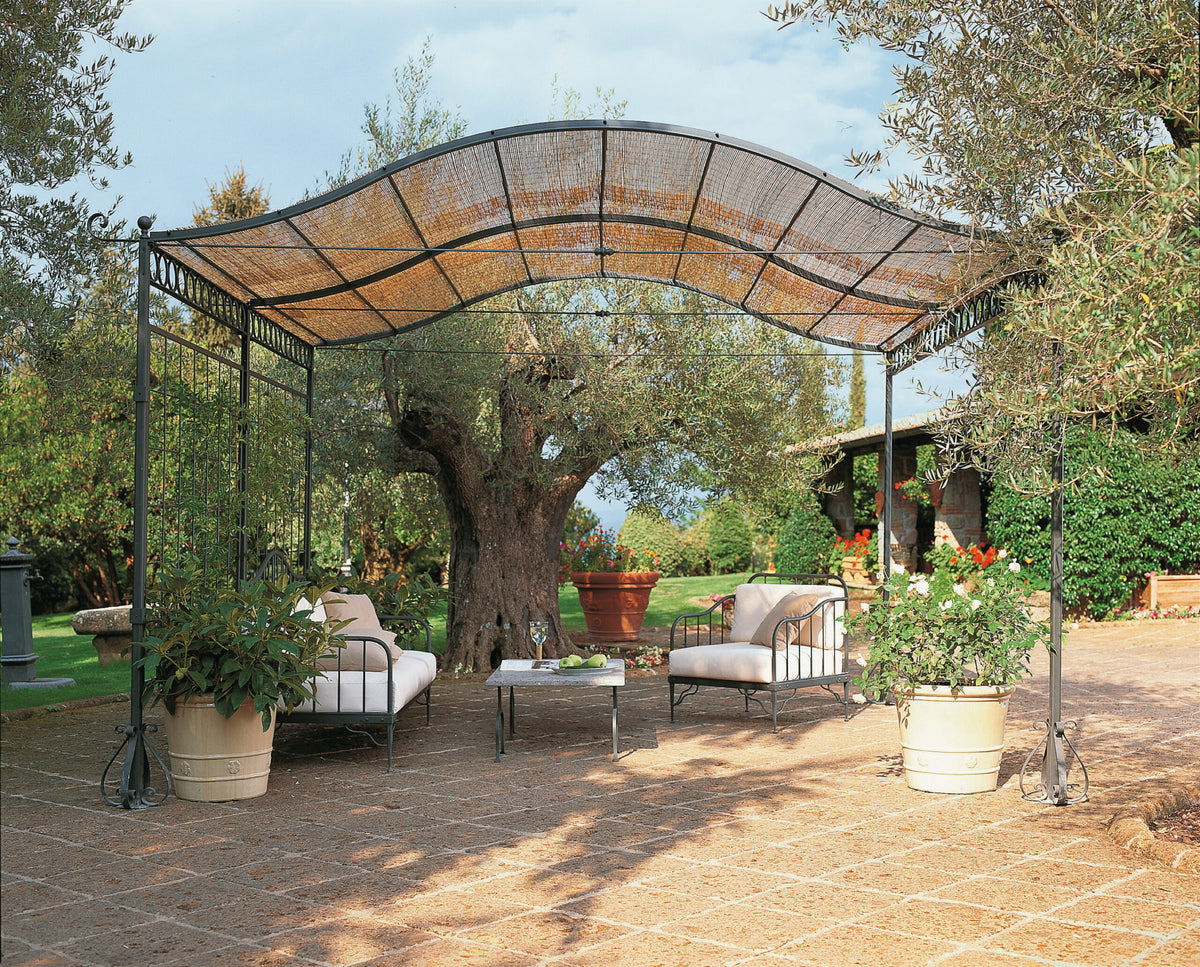 Eine freistehende, gebogene Pergola Solaire mit Metallrahmen und geflochtenem Dach spendet Schatten über einem Sitzbereich im Freien mit gepolsterten Stühlen und einem kleinen Tisch. Topfpflanzen schmücken den Raum, und im Hintergrund sind ein großer Baum und üppiges Grün zu sehen, die eine ruhige Gartenumgebung schaffen.