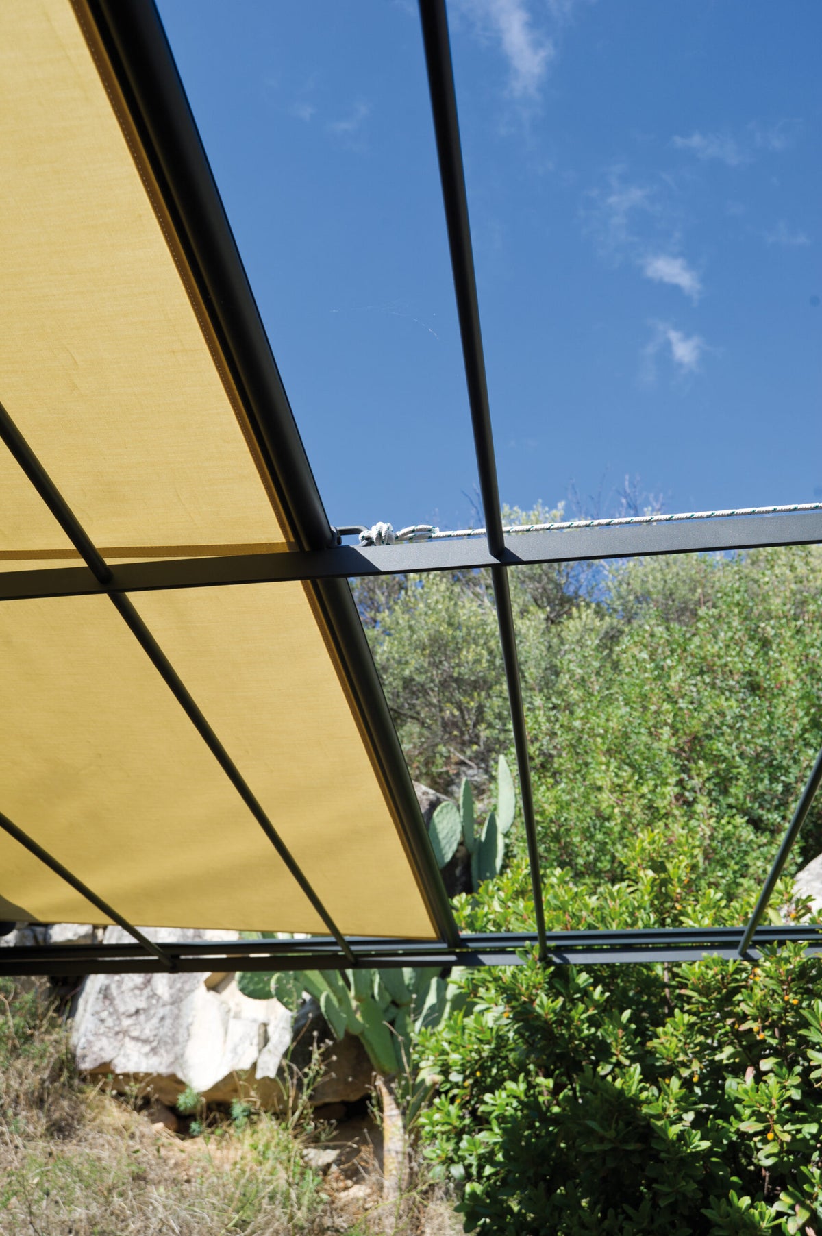 Von unten sieht man eine teilweise überdachte Gebogene Anbaupergola Solaire mit beigen Stoffschirmen, die den Blick auf einen blauen Himmel und üppiges Grün, darunter einen Kaktus und Büsche, einrahmt. Das Metallgerüst der Pergola bildet einen Kontrast zur natürlichen Umgebung.