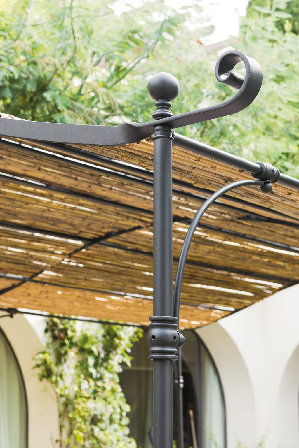 Eine Nahaufnahme eines schwarzen Metallpfostens an der Gebogenen Anbaupergola Solaire mit einem eleganten Schnörkeldesign an der Spitze. Hinter dem Pfosten befindet sich ein Lamellendach aus dünnen Holzstreifen, das teilweise von Bäumen beschattet wird. Die Solaire-Struktur wertet die Szene mit gewölbten Fenstern und üppigem grünem Laub im Hintergrund auf.