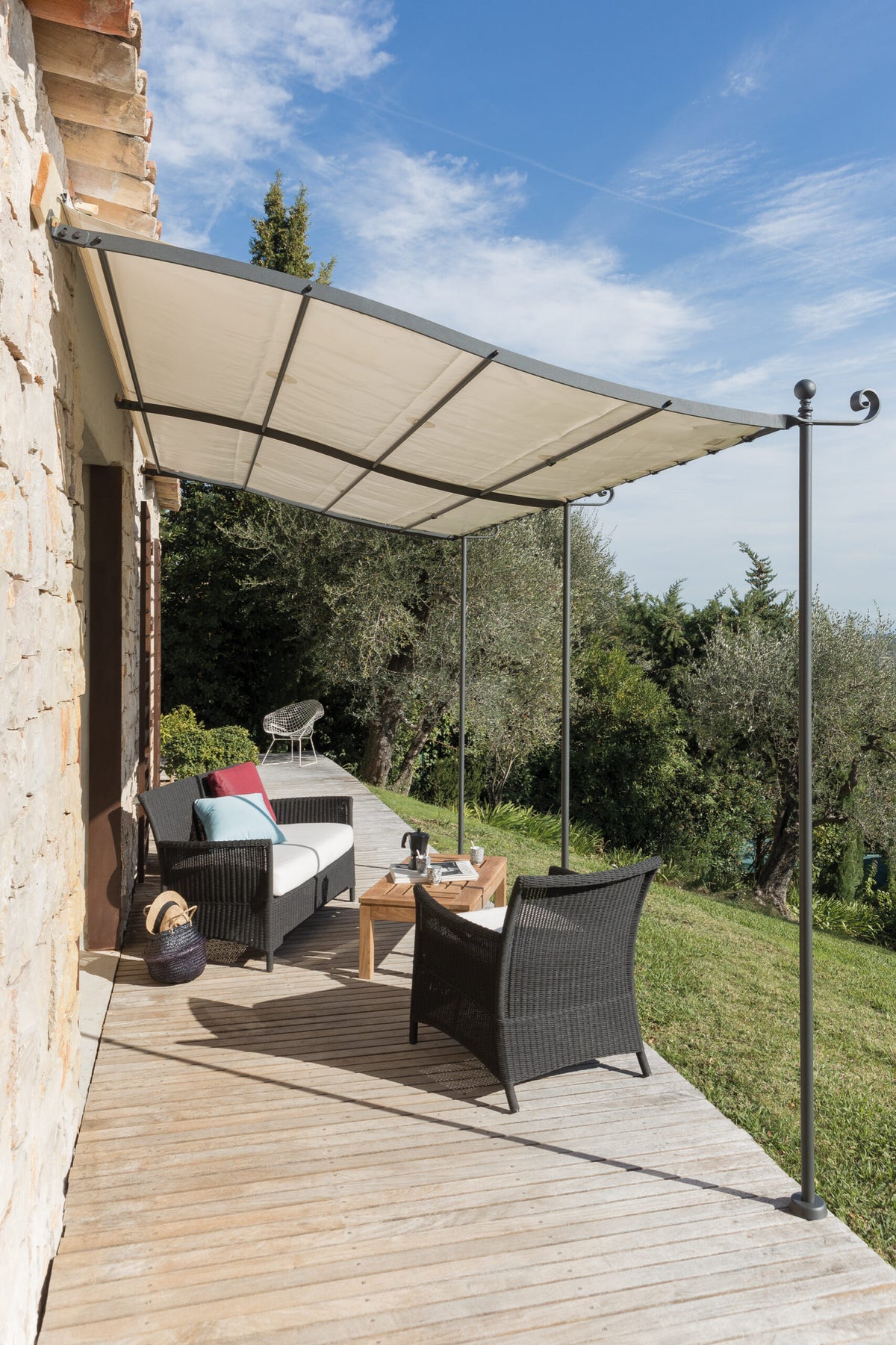 Eine gemütliche Außenterrasse mit Korbmöbeln, darunter ein Sofa und zwei Stühle um einen kleinen Couchtisch mit einer Tasse und einem Buch darauf. Die Terrasse hat einen Holzdeckboden und wird von der Gebogenen Anbaupergola Solaire beschattet. Im Hintergrund sind üppiges grünes Gras und Bäume unter klarem Himmel zu sehen.