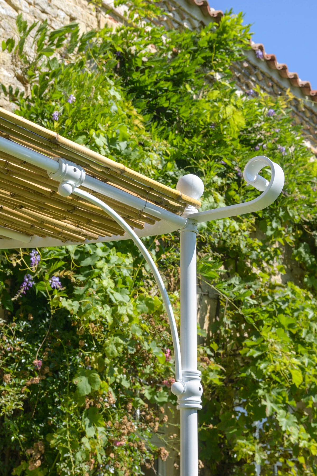 Nahaufnahme eines weißen, dekorativen Metallpfostens, der einen Bambusdachabschnitt auf der Lineare Anbaupergola Solaire stützt, vor dem Hintergrund einer üppigen grünen, mit Weinreben bewachsenen Steinmauer unter einem klaren blauen Himmel. Der Pfosten weist oben ein aufwendiges Schnörkelwerk auf.
