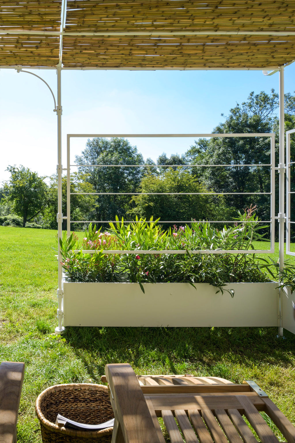 Eine malerische Aussicht auf einen Außenbereich zeigt eine Lineare Anbaupergola Solaire aus Bambus, die Schatten spendet. Darunter trennt ein weißes Metallgeländer im Lineare-Design ein mit grünen Pflanzen und Blumen gefülltes Gartenbeet von einer Rasenfläche. Im Vordergrund sind unter dem klaren Himmel ein Teil eines Holzstuhls und ein Weidenkorb zu sehen.