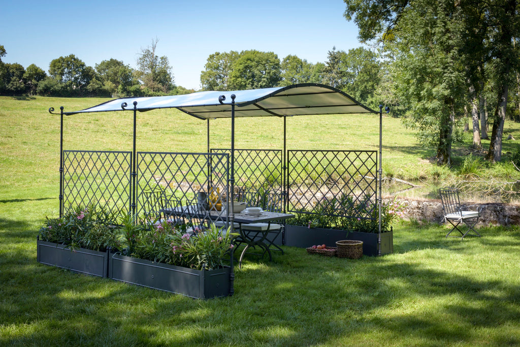 Eine freistehende, gebogene Pergola Solaire mit weißem Dach steht auf einer Wiese. Darunter stehen ein Tisch und Stühle, umgeben von Blumentöpfen. Im Hintergrund sind Bäume und Hecken zu sehen, und auf der Wiese außerhalb der Pergola stehen ein weiterer Stuhl und ein Korb.