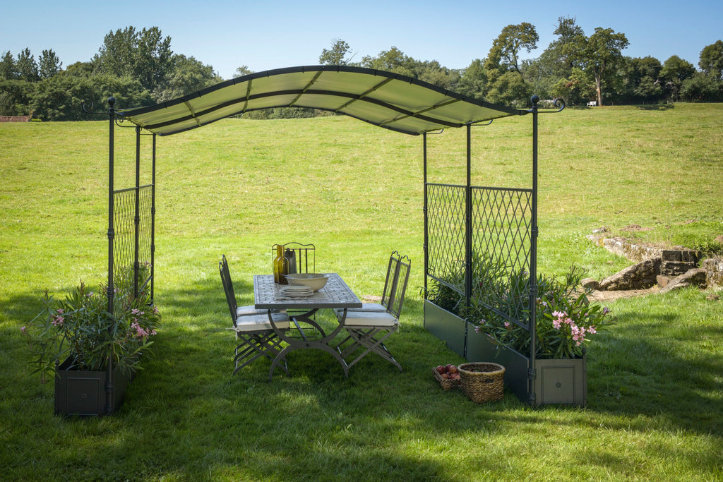 Ein Terrassen-Essset mit einem rechteckigen Tisch, vier Stühlen und der freistehenden, gebogenen Pergola Solaire mit geschwungenem Baldachin steht draußen auf einem üppigen grünen Rasen. Die Einrichtung ist von Pflanzkübeln mit blühenden Blumen umgeben, und im Hintergrund sind Bäume unter einem klaren Himmel zu sehen.