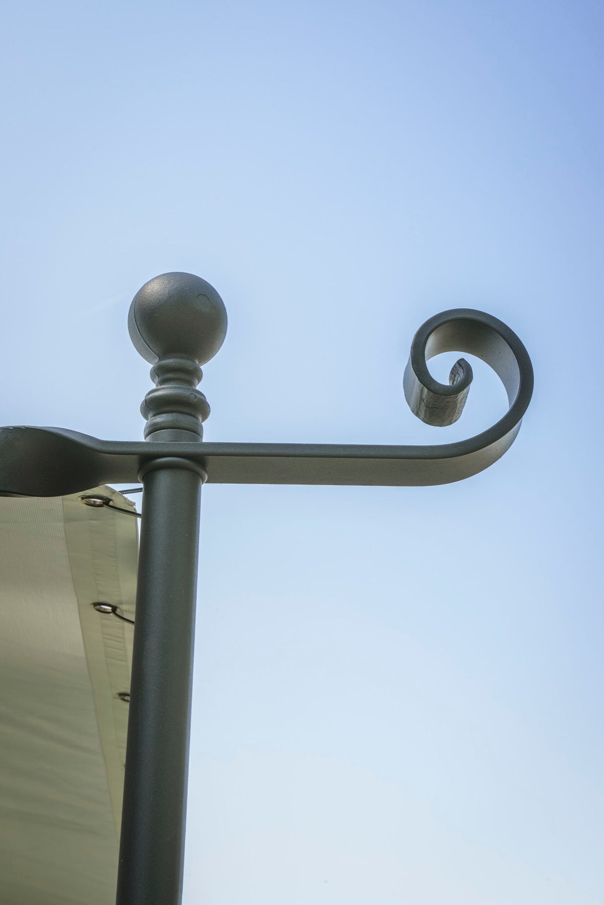 Nahaufnahme einer dekorativen Eisenstange mit einer Kugelspitze und einem Schnörkelmuster an der Spitze. Die Stange trägt ein gespanntes Stoffdach, das denen der linearen Anbaupergola Solaire-Strukturen ähnelt, die links vor einem klaren blauen Himmel zu sehen sind.