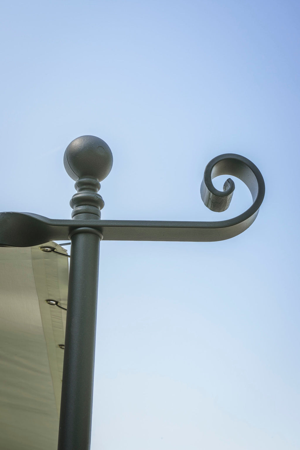 Nahaufnahme einer dekorativen schwarzen Metallstange mit Spiralmuster an einem sonnigen Tag. Unten links im Bild ist ein Teil eines befestigten grünen Stoffes zu sehen, möglicherweise ein Baldachin oder eine Markise, der mit Metallverschlüssen befestigt ist. Diese **Gebogene Anbaupergola Solaire** hat einen klaren, blauen Himmel im Hintergrund.