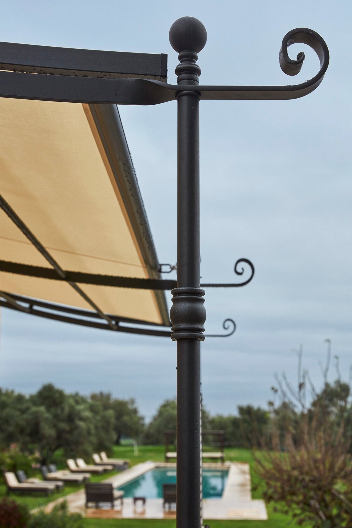 Eine Nahaufnahme einer Metallstütze der Lineare Anbaupergola Solaire mit beigem Baldachin und dekorativem Spiraldesign. Im Hintergrund ist ein ruhiger Außenbereich mit Liegestühlen, Bäumen und einem langen rechteckigen Swimmingpool unter einem bewölkten Himmel zu sehen.