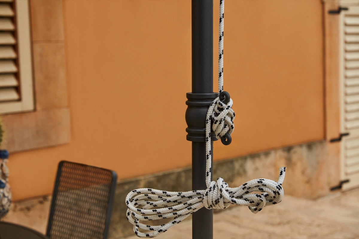 Um eine schwarze Stange ist ein dickes weißes Seil mit schwarzen Markierungen kunstvoll gebunden, vor dem Hintergrund eines Gebäudes mit Fensterläden und einem gefliesten Boden. Auf der linken Seite ist teilweise ein schwarzer Stuhl mit Netzrücken zu sehen, der das schlanke Design einer Lineare Anbaupergola Solaire in der Nähe perfekt ergänzt.