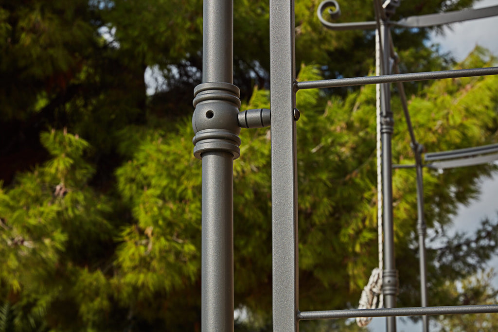 Nahaufnahme einer Metalltischlereikonstruktion mit zylindrischen Verbindungsstücken und Bolzen vor einer Kulisse üppiger grüner Bäume, die sich nahtlos in das schlanke Design der Lineare Anbaupergola Solaire einfügt.