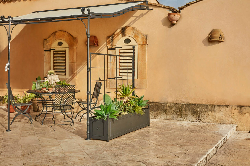 Eine gemütliche Außenterrasse mit einem Metalltisch und Stühlen unter einer linearen Anbaupergola Solaire. Die Terrasse ist mit verschiedenen Topfpflanzen und Sukkulenten geschmückt, die durch integrierte Lichter beleuchtet werden. Der Hintergrund besteht aus einer rustikalen beigefarbenen Wand mit zwei Bogenfenstern und einigen dekorativen Töpferwaren.