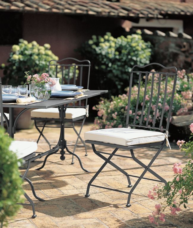 Eine bezaubernde Außenterrasse, die an die Toskana erinnert, verfügt über einen Tisch mit Gläsern, Geschirr und einer Vase mit rosa Blumen. Klappbare Stühle aus Metall der Serie „Toscana“ mit weißen Kissen umgeben den Tisch. Üppiges Grün und blühende Blumen schmücken den Hintergrund und schaffen eine ruhige und einladende Atmosphäre.