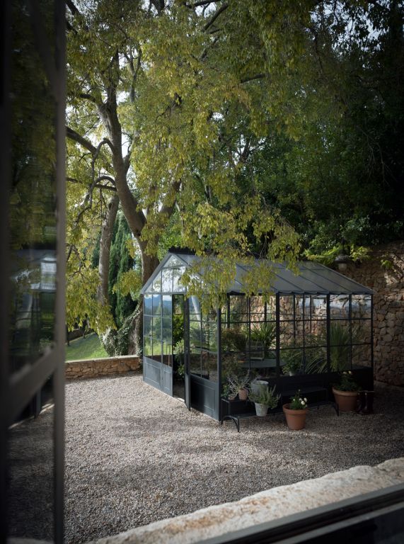 Eine kleine freistehende Orangerie mit Glaspaneelen steht auf einer Kiesfläche im Schatten eines großen Baumes. Mehrere Blumentöpfe stehen außerhalb dieses eleganten Gewächshauses. Steinmauern und dichtes Grün umgeben die Szene und schaffen eine ruhige Gartenumgebung.