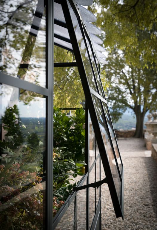 Eine moderne freistehende Orangerie mit einem teilweise geöffneten Fenster, das den Blick auf üppige grüne Pflanzen im Inneren freigibt. Der schlanke Metallrahmen und die Glasplatten unterstreichen das zeitgenössische Design. Im Hintergrund führt ein von Bäumen gesäumter Kieselweg zu einer malerischen Fernsicht.